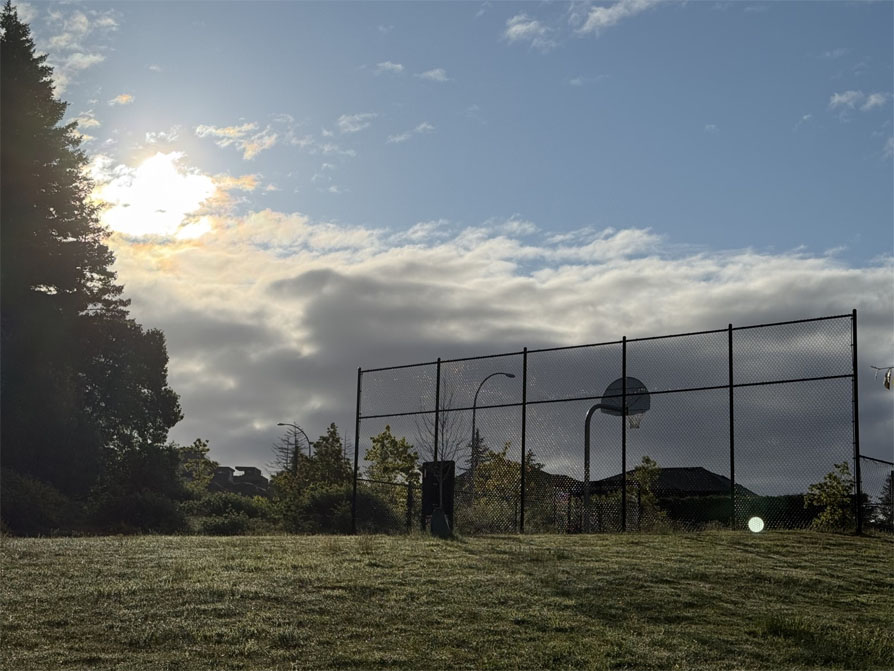 Photo the sun rising above some clouds over a shaded basketball court.