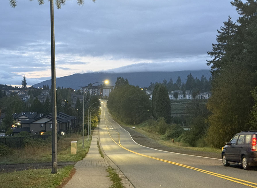 Photo of a quiet road leading down a hill and up in the distance.
	A single dark-coloured car just enters the photo on the right side.