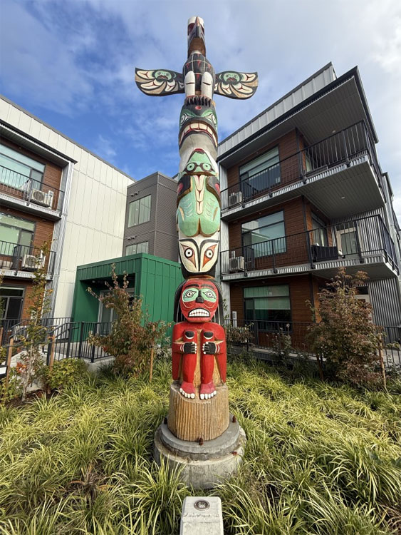 Photo of a brightly coloured totem pole surrounded by low shrubs, placed in front of an apartment-style building
	as a local art feature.