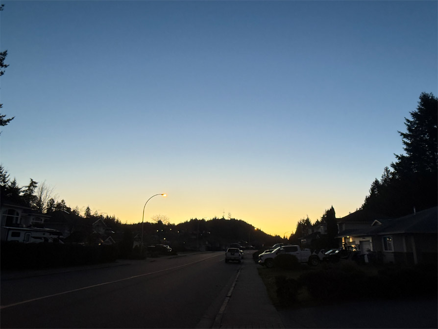 Image shows the line of a neighbourhood and hill in darkness; the sun rising behind makes a sharp yellow line over the hill.