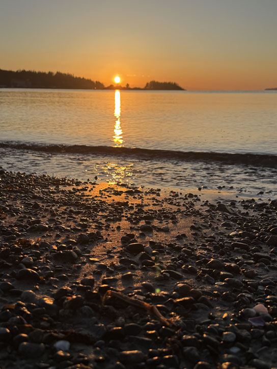 Photo of a sunset, the sun making a vertical bright line across the water down to sand and rocks that glow and glitter.